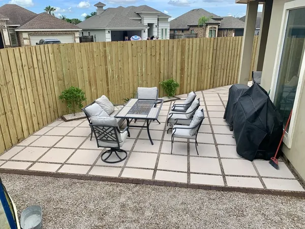 A back patio of large pale square pavers set with wide open joints, with a table, chairs and a covered grill against a wooden fence