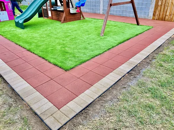 A rectangle of artificial turf edged by a red band and a tan paver band, with a wooden swing set and a green slide standing on it