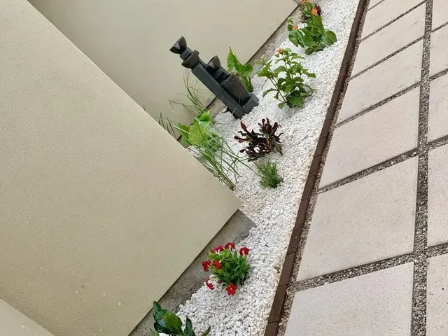 A narrow strip of white gravel between a stucco wall and a run of large pavers, with a sprinkler head standing in it
