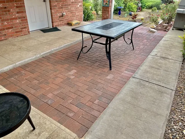 A red and tan paver patio beside a brick house, with a patio table on it and a fire bowl in the foreground