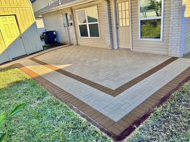 A back patio of tan pavers laid in herringbone, with a dark brown band running around it inside a lighter outer edge