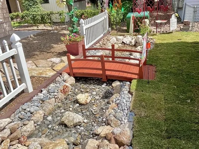 A dry creek bed of river rock and stones running through a lawn, with a small red arched wooden bridge over it