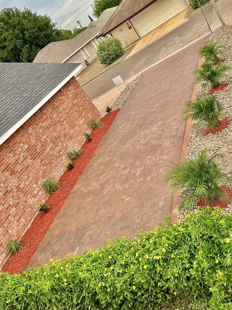 A red and tan paver run along the side of a brick house, with a narrow strip of red mulch planted with small shrubs
