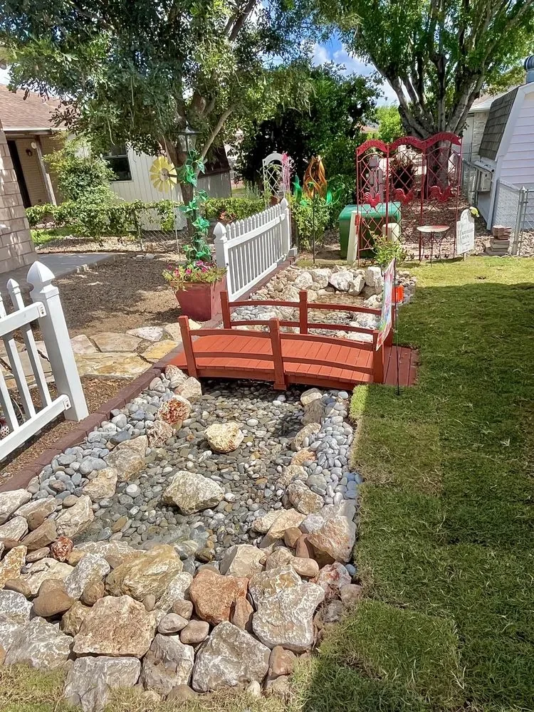 A dry creek bed of river rock running through a lawn, with a small red arched wooden bridge over it and a white bench behind