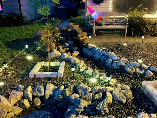 A rock bed at night, winding between boulder borders with low path lights along it and a tree in a stone-edged square